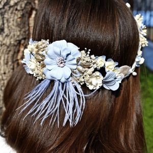 Corona de flores azul pastel y blanco con hojas decorativas doradas y plateadas, detalles florales beige y plumas azules.